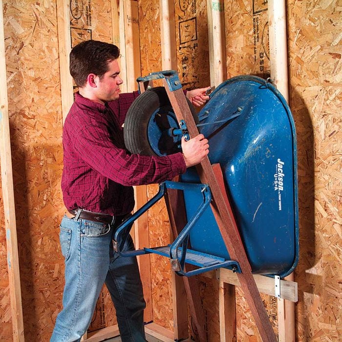 A person in a red shirt is lifting a blue wheelbarrow onto a wooden plank in a shed or an unfinished room with exposed plywood walls and wooden studs. The space appears to be under construction or being used for storage.