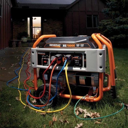 A Generac XG7000E generator is shown on a lawn with multiple colored cords plugged into it. The generator is positioned near a house with a lit doorway in the background, suggesting it is being used to power the home.